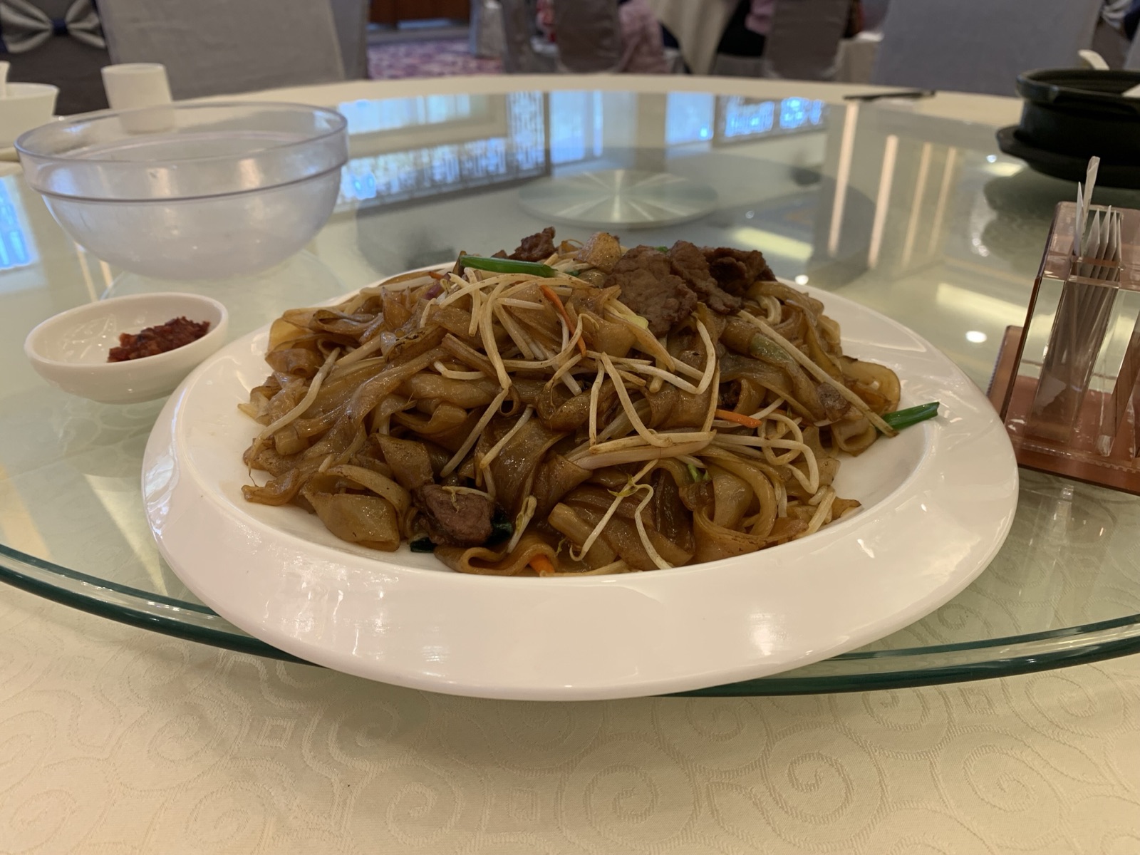 Beef chow fun on a banquet lazy-susan table with chilli sauce alongside